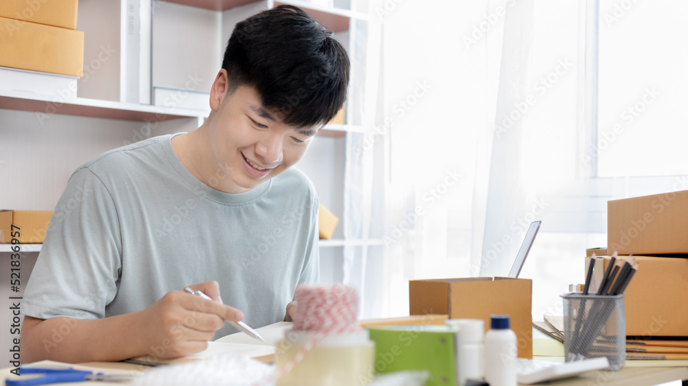 Young Asian man is writing down the customer's details and addresses on the notebook or box in order to prepare for shipping according to the information, Packing box, Sell online, freelance working. Young Asian man is writing down the customer's details and addresses on the notebook or box in order to prepare for shipping according to the information, Packing box, Sell online, freelance working.