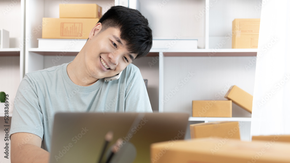 Man talking on the phone and typing on the computer chatting with customers who place orders online, Packaging on background, Sell online, Small business owner, Online shopping SME entrepreneur. Man talking on the phone and typing on the computer chatting with customers who place orders online, Packaging on background, Sell online, Small business owner, Online shopping SME entrepreneur.