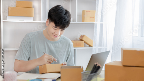 Young Asian man is writing down the customer's details and addresses on the notebook or box in order to prepare for shipping according to the information, Packing box, Sell online, freelance working.