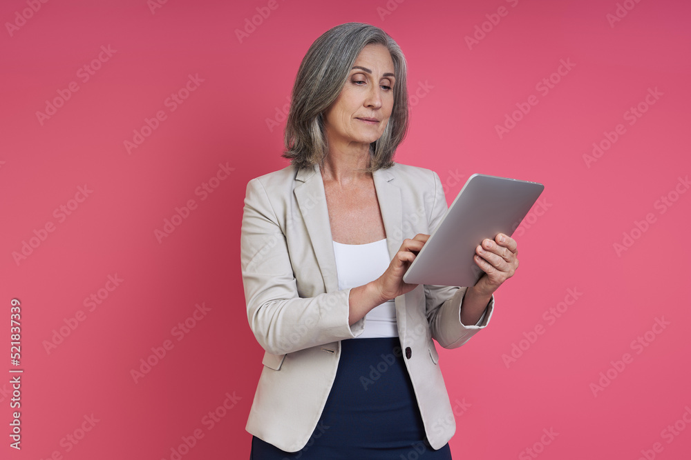 Confident senior woman in formalwear using digital tablet while standing against pink background