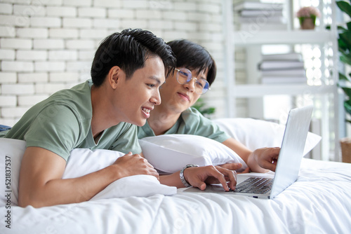 Happy Asian gay couple using laptop and relaxing at home on bed, LGBTQ concept.