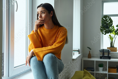 Worried young woman looking through a window while sitting on the window sill © gstockstudio