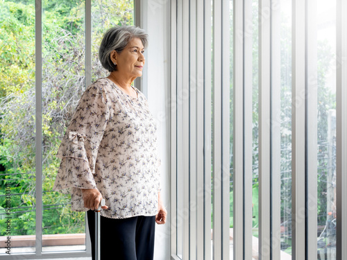 Asian senior woman  white hair standing with a cane and looking out the glass window  indoors with copy space. Elderly lady patient using walking cane. Strong health  medical care concepts.