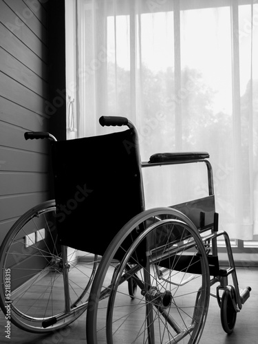 The back view of empty black wheelchair near wall with white curtain at glass window in the living room vertical style, waiting for patient services. Lonely wheelchair with nobody in hospital room.