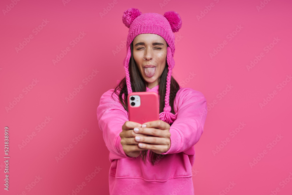 Playful young woman in funky hat holding smart phone and grimacing against pink background Playful young woman in funky hat holding smart phone and grimacing against pink background