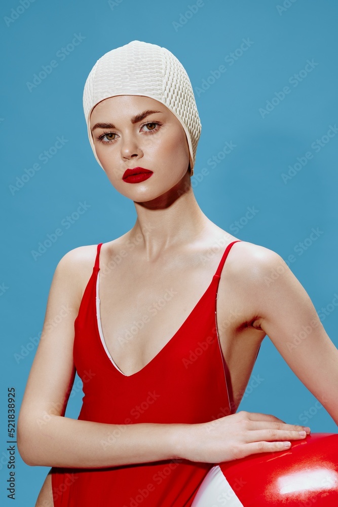 A concentrated beautiful lady in a swimming cap, a red swimsuit with a ball on a blue background looks at the camera. Conceptual fashion A concentrated beautiful lady in a swimming cap, a red swimsuit with a ball on a blue background looks at the camera. Conceptual fashion