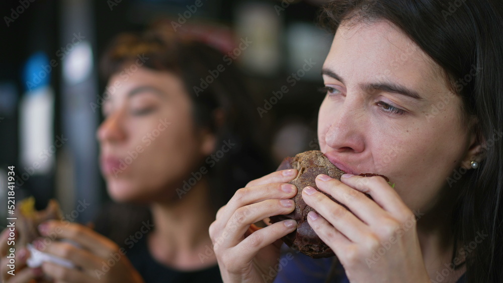 Two people eating burgers. Young women taking a bite of cheeseburgers. Female friends eating fast food lunch Two people eating burgers. Young women taking a bite of cheeseburgers. Female friends eating fast food lunch