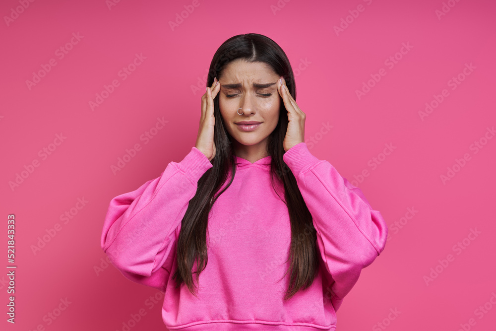Fototapeta premium Frustrated woman touching head with hands and keeping eyes closed against pink background