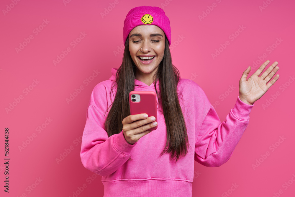 Beautiful young woman in hooded shirt and funky hat holding smart phone against pink background Beautiful young woman in hooded shirt and funky hat holding smart phone against pink background