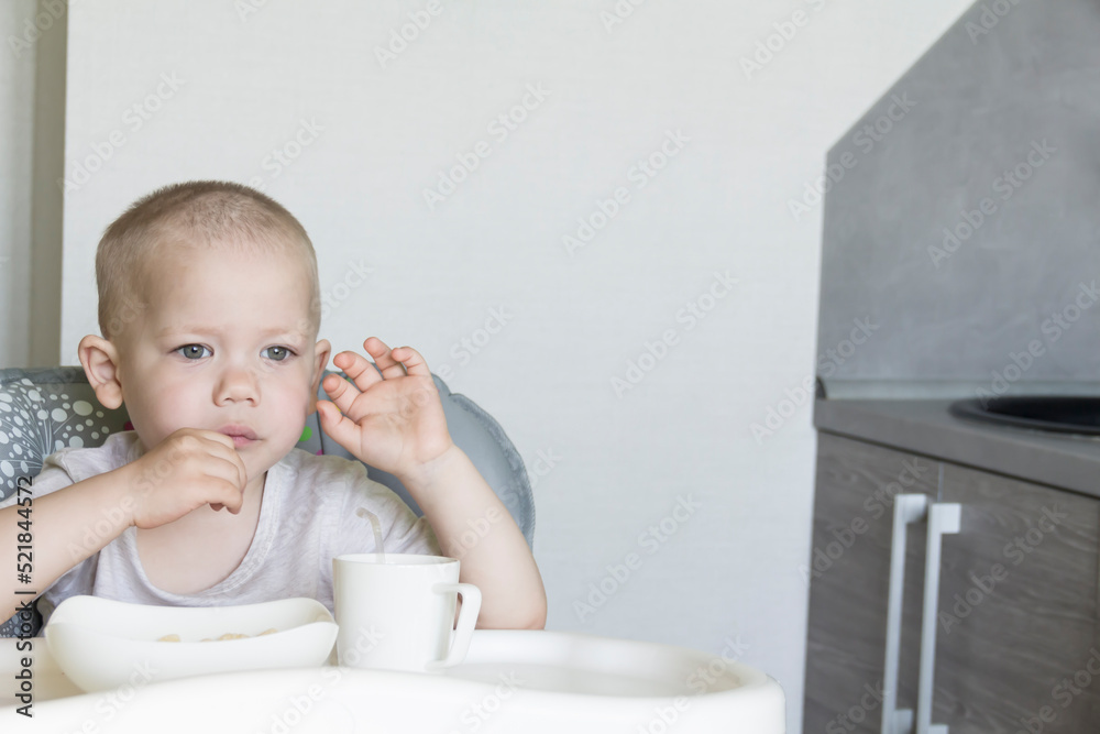 A little boy with a short haircut independently and appetitiously eats food and drinks juice from a tube in a child's chair. A little boy with a short haircut independently and appetitiously eats food and drinks juice from a tube in a child's chair.