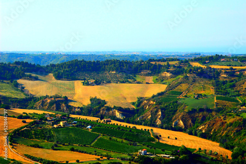 Rural view in Acquaviva Picena with a rugged Marche landscape consisting of green vegetation, yellow meadows, peculiar ravines and scattered houses and buildings under a hazy azure sky