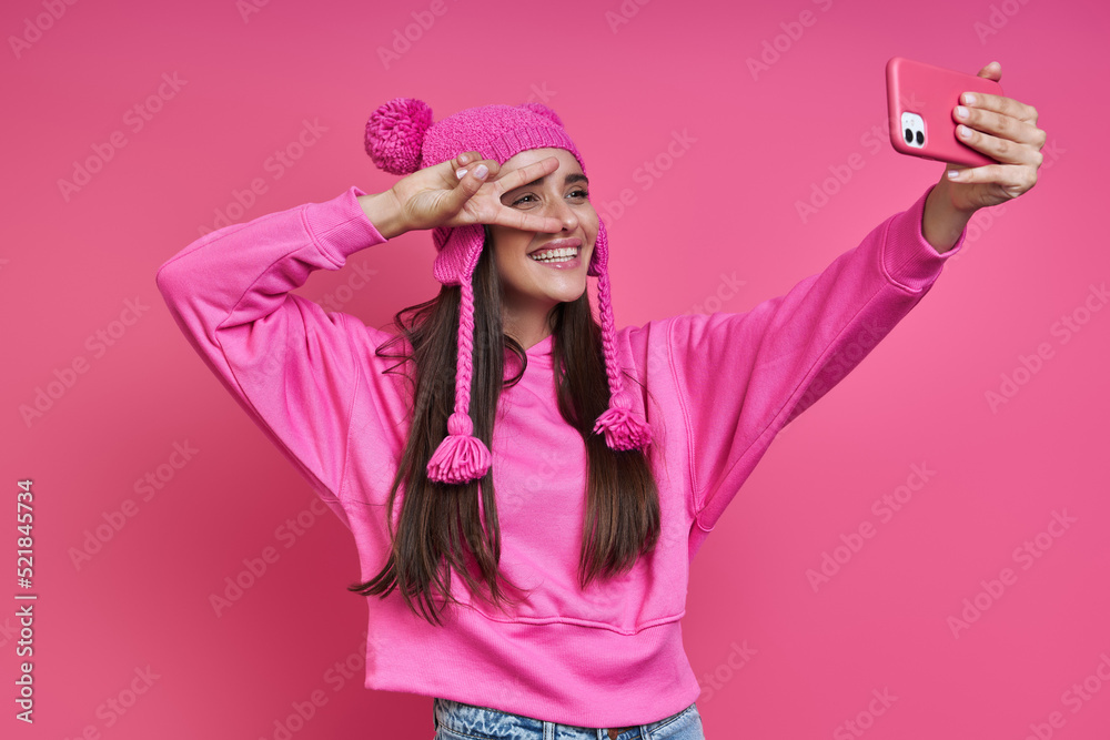 Beautiful young woman in funky hat making selfie and gesturing against pink background Beautiful young woman in funky hat making selfie and gesturing against pink background