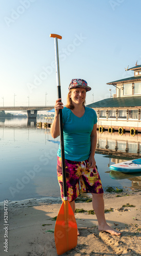 A young girl wearing a full cap, blue T-shirt and board short stands with a paddle