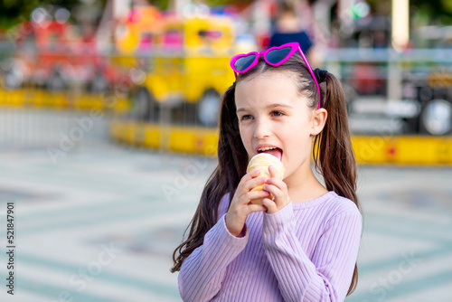 portrait of a child girl in an amusement park in the summer eating ice cream near the carousels and smiling with happiness in sunglasses, the concept of weekends and school holidays