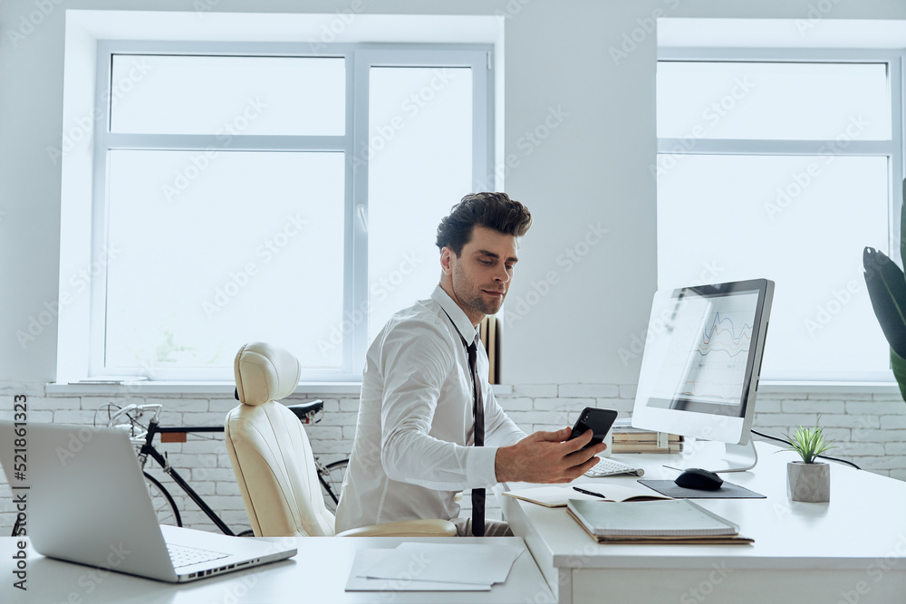 Confident man in shirt and tie looking at his smart phone while sitting in office Confident man in shirt and tie looking at his smart phone while sitting in office