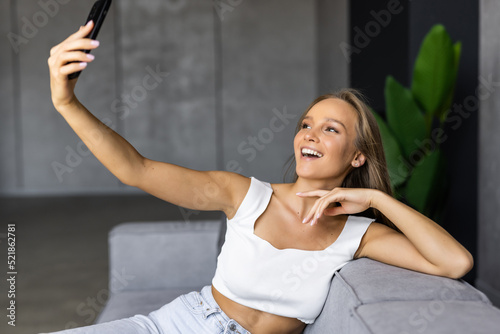Pretty smiling young woman sitting at sofa, posing a smile and taking a selfie with phone.