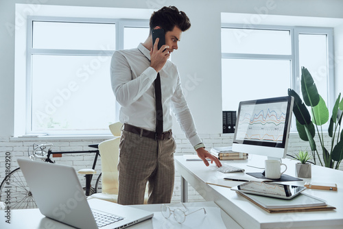 Confident young man looking at computer while talking on mobile phone in office © gstockstudio