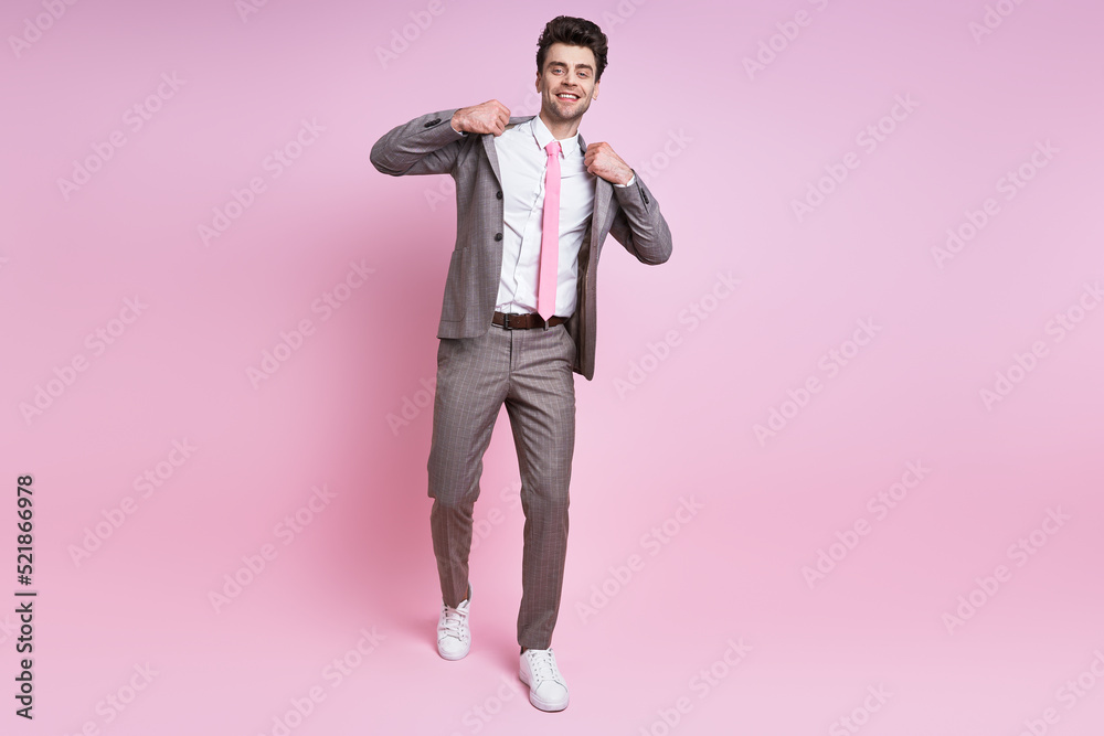 Cheerful young man in full suit adjusting his jacket while standing against pink background Cheerful young man in full suit adjusting his jacket while standing against pink background