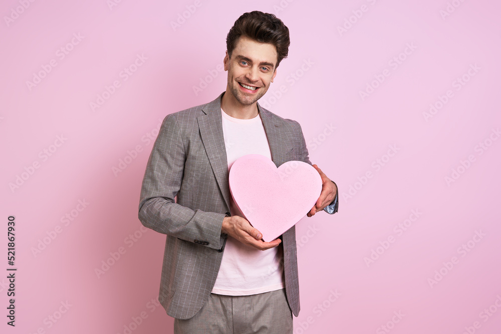 Happy young man in suit holding paper heart while standing against pink background Happy young man in suit holding paper heart while standing against pink background
