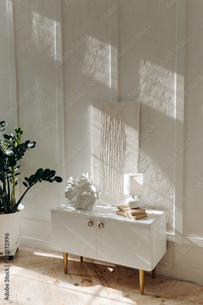 Stylish white bedside table with decor, lamp and white abstract gypsum painting against a textured white wall. Potted plant at home. Sunbeams in the interior. Nobody. Stylish white bedside table with decor, lamp and white abstract gypsum painting against a textured white wall. Potted plant at home. Sunbeams in the interior. Nobody.