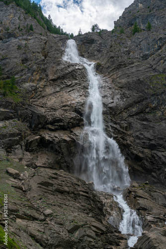 grosser Wasserfall der Engstligen in Adelboden. Das Wasser fett über eine hohe Felswand auf Geröll, Steine und Felsen. Am Ufer hat es Gras, Blumen und Bäume