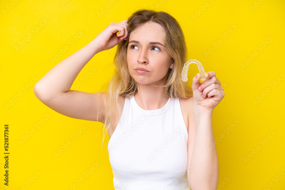 Blonde English young girl holding invisible braces isolated on yellow background having doubts and with confuse face expression Blonde English young girl holding invisible braces isolated on yellow background having doubts and with confuse face expression