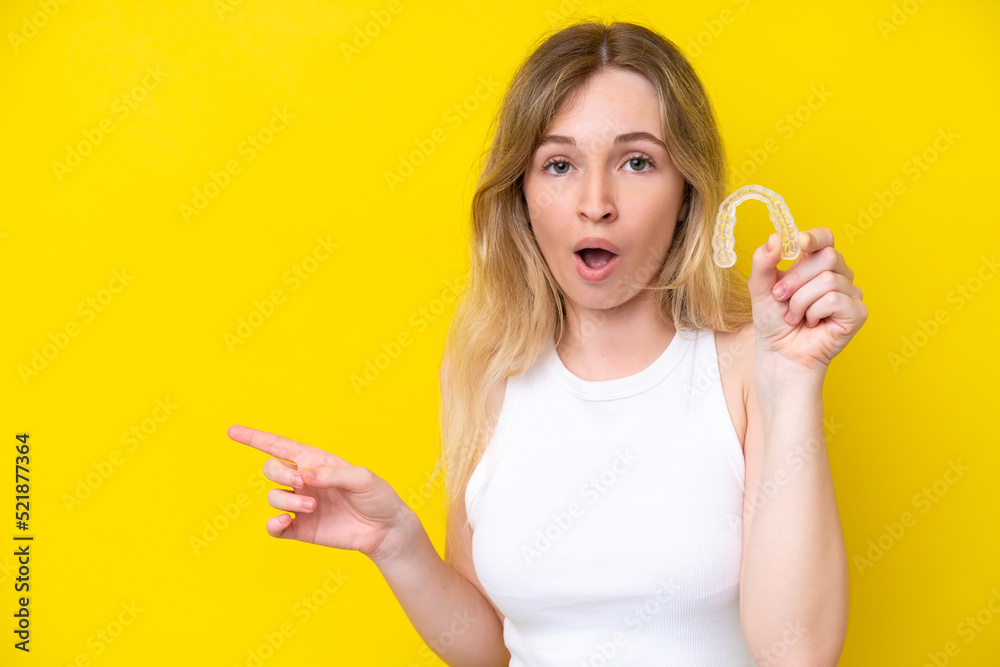 Blonde English young girl holding invisible braces isolated on yellow background surprised and pointing side Blonde English young girl holding invisible braces isolated on yellow background surprised and pointing side