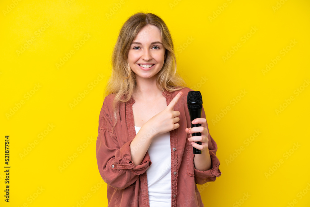 Young singer caucasian woman picking up a microphone isolated on yellow background pointing to the side to present a product Young singer caucasian woman picking up a microphone isolated on yellow background pointing to the side to present a product