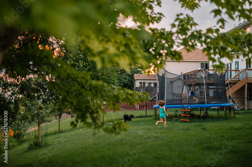 A girl runs across a green suburban back yard to a trampoline that her brother is doing a flip on at sunset