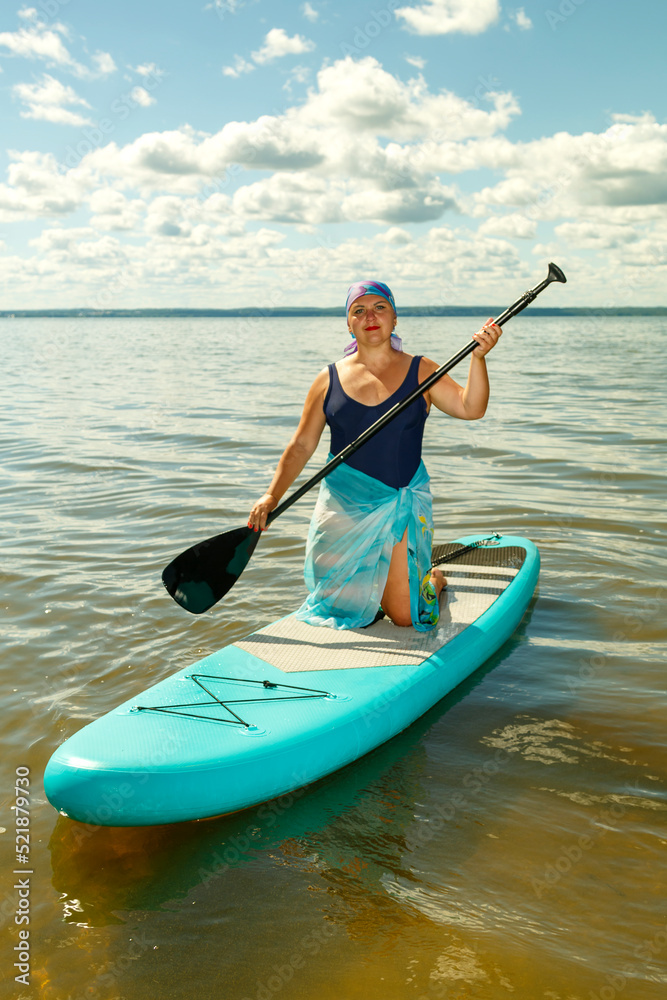 A Jewish woman in a pareo and a headdress stands on a SUP board with an oar on the lake.
