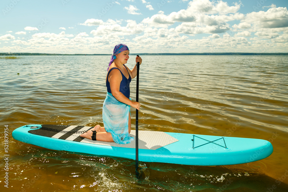 A Jewish woman in a closed swimsuit, a pareo and a headdress on her knees floats on a SUP board with an oar.