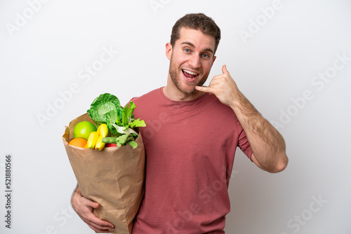 Young caucasian man holding a grocery shopping bag isolated on white background making phone gesture. Call me back sign