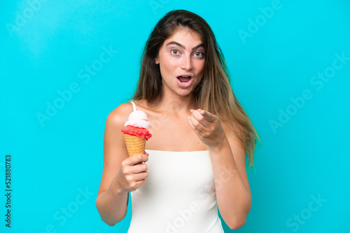Young woman in swimsuit holding an ice cream isolated on blue background surprised and pointing front
