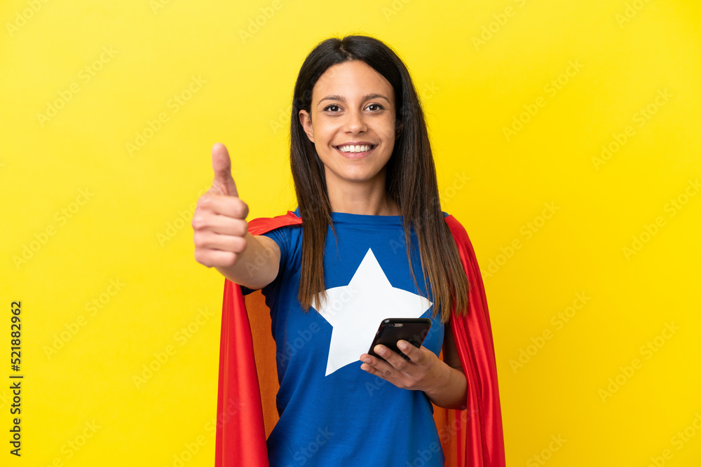 Super Hero woman isolated on yellow background using mobile phone while doing thumbs up Super Hero woman isolated on yellow background using mobile phone while doing thumbs up