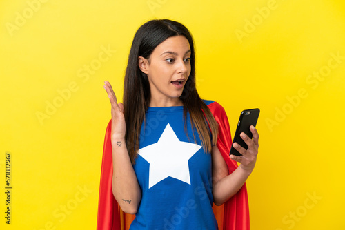 Super Hero woman isolated on yellow background looking at the camera while using the mobile with surprised expression