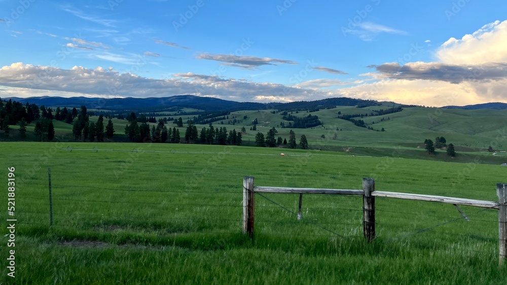 Wooden gate and barbed wire fencing in foreground, with grass land and pine-covered hills beyond. Overcast, dark, with a few clouds overhead. Montana, USA