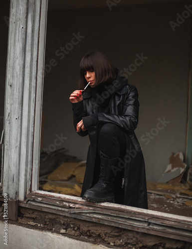 Woman in long leather coat at window of destroyed apartment building thoughtfully smokes cigarette