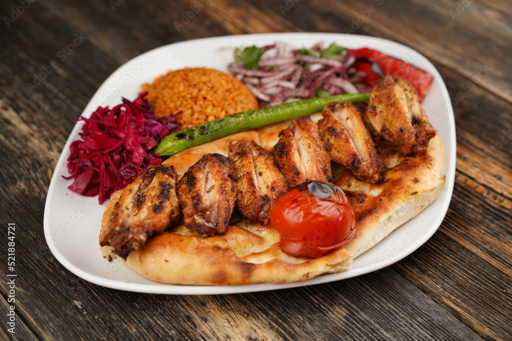 Grilled Turkish chicken wings served with pita, garnish, onions, bulgur, roasted green pepper, and roasted tomato on white plate on wooden background. Served special restaurant presentation Grilled Turkish chicken wings served with pita, garnish, onions, bulgur, roasted green pepper, and roasted tomato on white plate on wooden background. Served special restaurant presentation