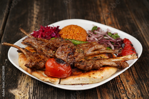 Grilled lamb chops on a white plate on wooden background. Grilled lamb with pita, garnish,  onions, bulgur, roasted green pepper, and roasted tomato. Turkish name: kuzu pirzola. 