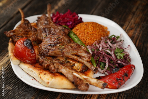 Grilled lamb chops on a white plate on wooden background. Grilled lamb with pita, garnish, onions, bulgur, roasted green pepper, and roasted tomato. Turkish name: kuzu pirzola. 