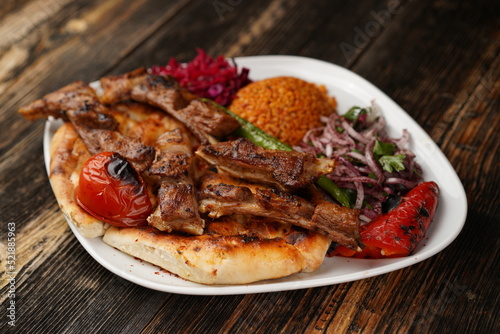 Grilled lamb chops on a white plate on wooden background. Grilled lamb with pita, garnish, onions, bulgur, roasted green pepper, and roasted tomato. Turkish name: kuzu pirzola. 