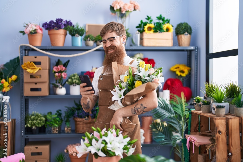 Young redhead man florist using smartphone holding flowers at flower shop