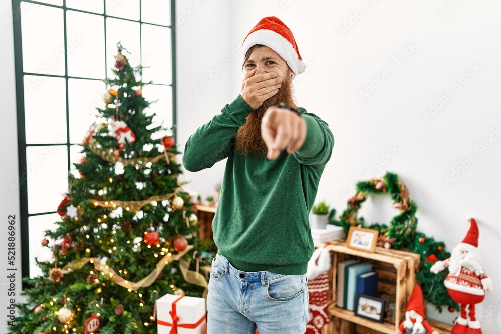 Redhead man with long beard wearing christmas hat by christmas tree laughing at you, pointing finger to the camera with hand over mouth, shame expression