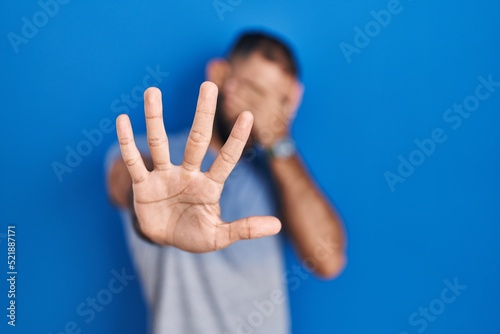 Middle east man with beard standing over blue background covering eyes with hands and doing stop gesture with sad and fear expression. embarrassed and negative concept.