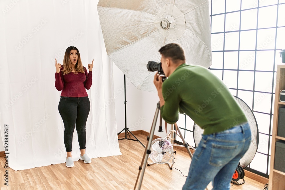 Young woman at photography studio amazed and surprised looking up and pointing with fingers and raised arms.