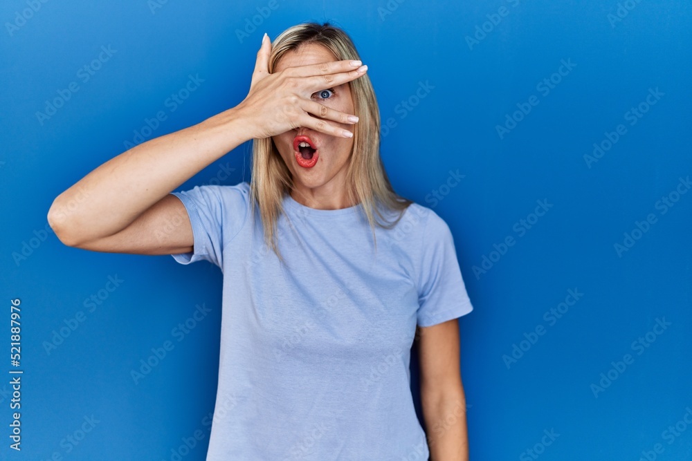 Beautiful blonde woman wearing casual t shirt over blue background peeking in shock covering face and eyes with hand, looking through fingers with embarrassed expression. Beautiful blonde woman wearing casual t shirt over blue background peeking in shock covering face and eyes with hand, looking through fingers with embarrassed expression.