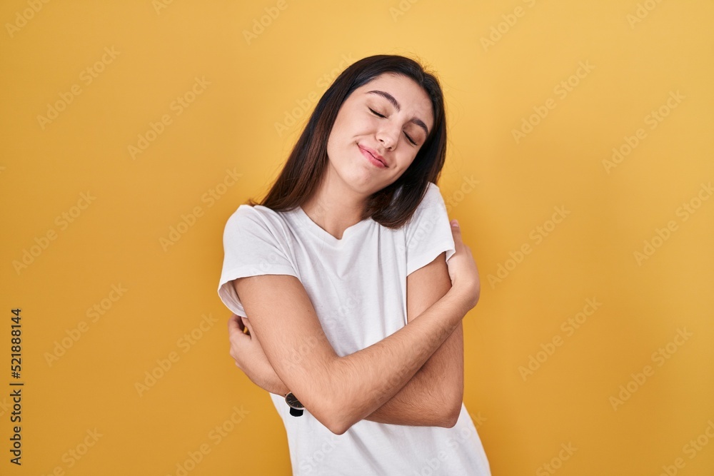 Young beautiful woman standing over yellow background hugging oneself happy and positive, smiling confident. self love and self care Young beautiful woman standing over yellow background hugging oneself happy and positive, smiling confident. self love and self care