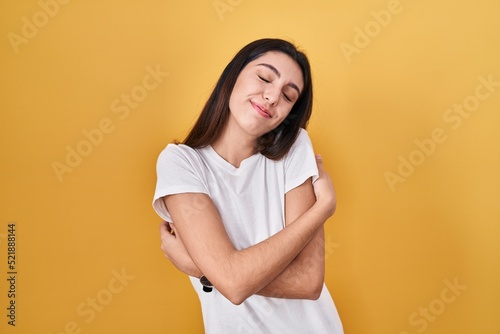 Young beautiful woman standing over yellow background hugging oneself happy and positive, smiling confident. self love and self care
