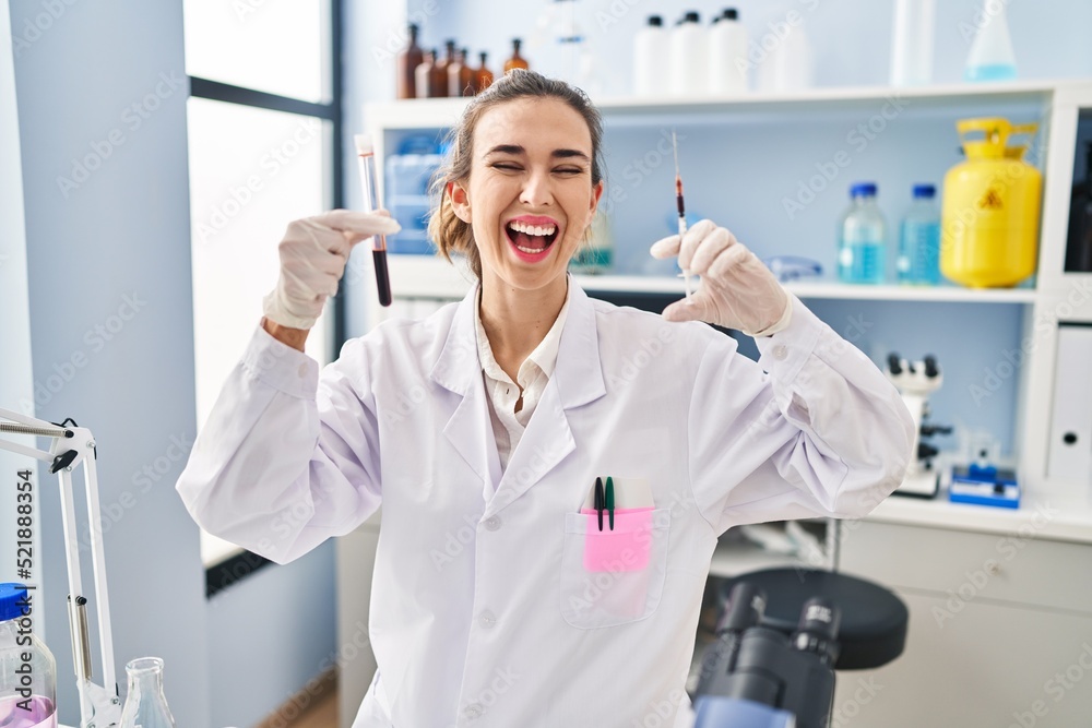 Young woman working at scientist laboratory holding blood sample smiling and laughing hard out loud because funny crazy joke. Young woman working at scientist laboratory holding blood sample smiling and laughing hard out loud because funny crazy joke.