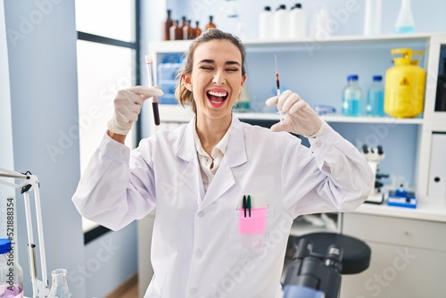 Young woman working at scientist laboratory holding blood sample smiling and laughing hard out loud because funny crazy joke.
